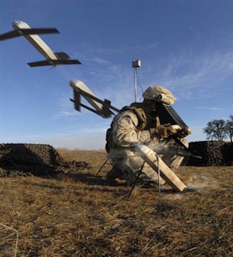 a soldier crouches on the ground peering into a black box as to small projectiles with wings are launched from tubes on either side of him