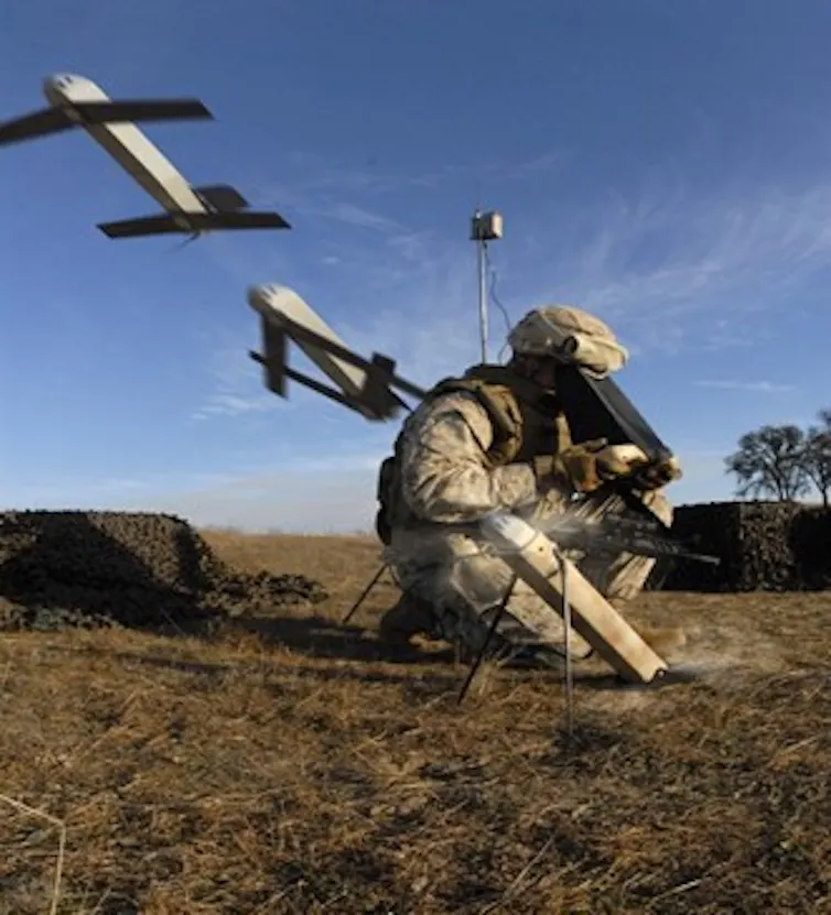 a soldier crouches on the ground peering into a black box as to small projectiles with wings are launched from tubes on either side of him