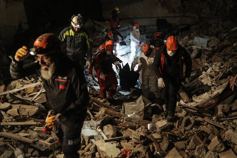Rescue workers in red helmets carry a body bag containing a body out of a collapsed building.