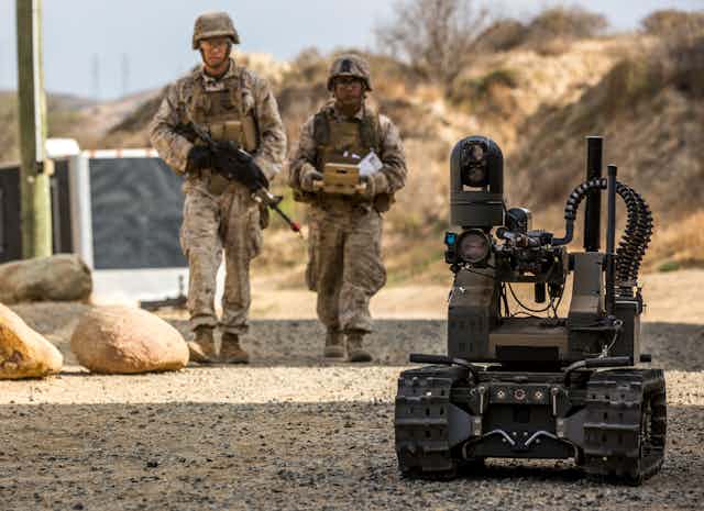 ttwo soldiers walk behind a machine gun mounted on a small treaded vehicle