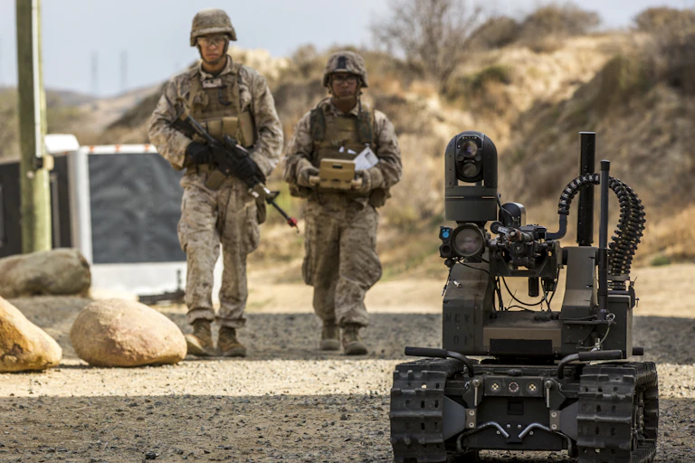 ttwo soldiers walk behind a machine gun mounted on a small treaded vehicle