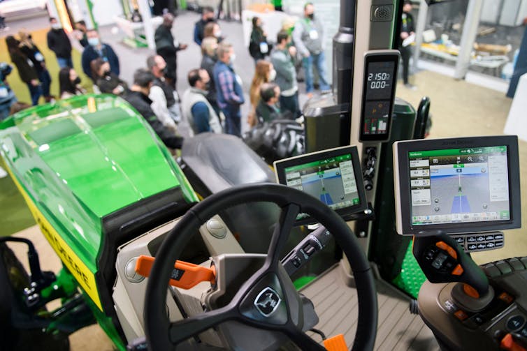 A tractor with several computer screens in the cab on the floor of a convention, with several people in the background.