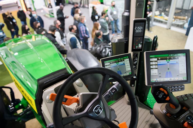 A tractor with several computer screens in the cab on the floor of a convention, with several people in the background.JD's Deal with US Farm Bureau raises hopes for Right to Repair