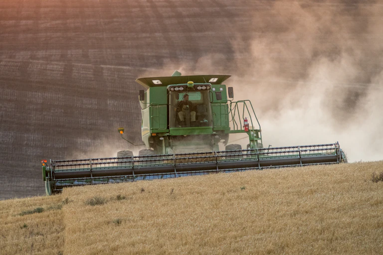A large combine moves over a hill, with an already cut field in the background. The farm can be seen in the cab.