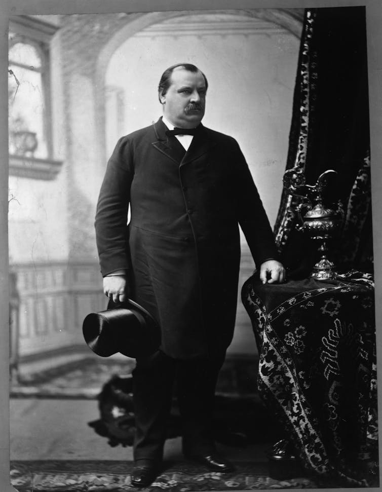 A suited man stands with a top hat in his right hand as his left hand rests on a side table dressed in a table cloth.