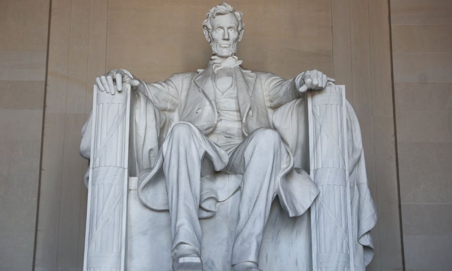 A large, white statue depicts a bearded man, sitting with both arms atop arm rests.