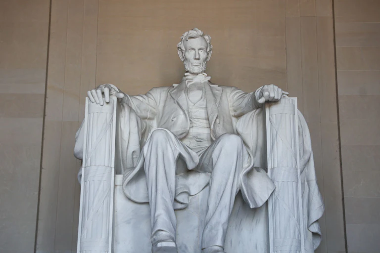 A large, white statue depicts a bearded man, sitting with both arms atop arm rests.