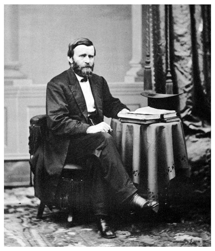 A bearded man, dressed in a suit, sits with his right leg crossed over his left. His left hand rests on a book, atop a side table.