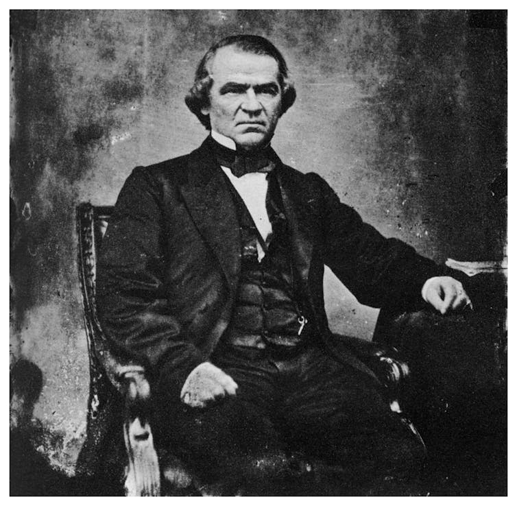 A suited man, with ear-length hair, sits with his left hand resting on a side table.