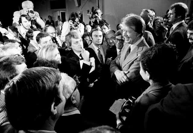 A smiling man in a light suit jacket and tie, surrounded by people who are listening to him.