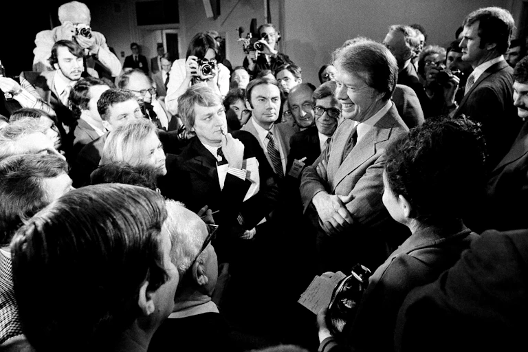 A smiling man in a light suit jacket and tie, surrounded by people who are listening to him.