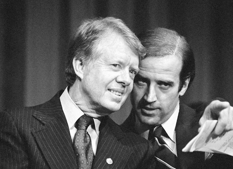 Two men in suits and ties, talking with their heads close.