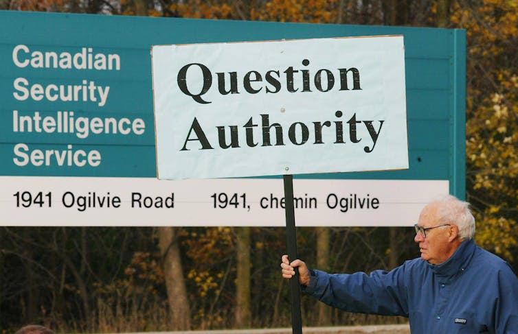 An elderly man carries a placard that reads: Question authority, in front of a roadside sign that says Canadian security Intelligence service