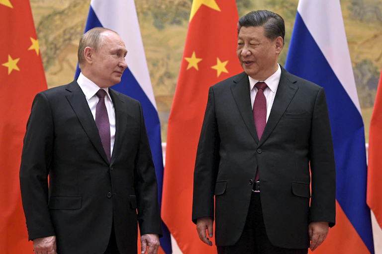 A white man and an asian man wearing suits stand in front of flags and look at each other warmly