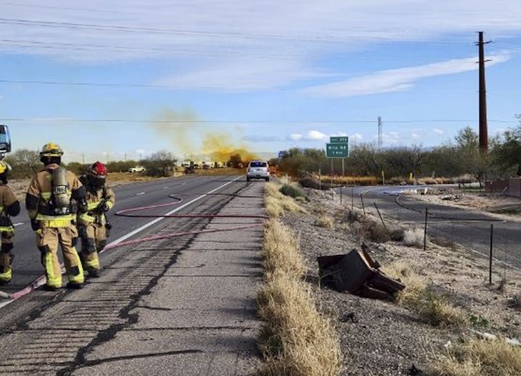 Los bomberos se paran en una carretera mientras un humo naranja se eleva en la distancia.