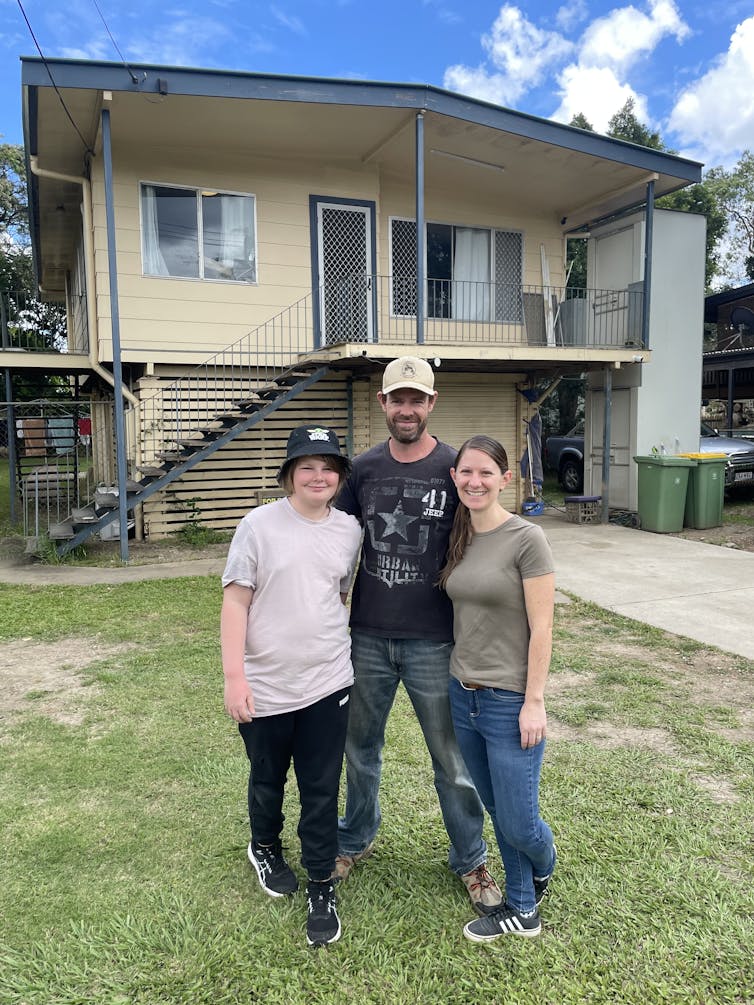 A family of three, Paul Harding in the centre wearing a cap, standing on the lawn in front of his home in Goodna, Qld.