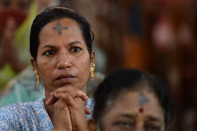 A woman with gold earrings and a black cross mark on her forehead folds her hands in prayer.