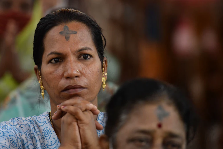 A woman with gold earrings and a black cross mark on her forehead folds her hands in prayer.