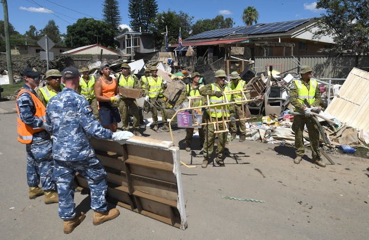 Members of the Australian Defence Force, wearing blue camo overalls, and local residents in hi-vis vests clean up flood-damaged properties in the suburb of Goodna in Ipswich.