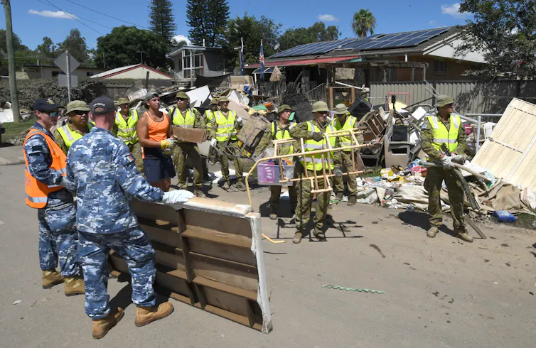 Members of the Australian Defence Force, wearing blue camo overalls, and local residents in hi-vis vests clean up flood-damaged properties in the suburb of Goodna in Ipswich.