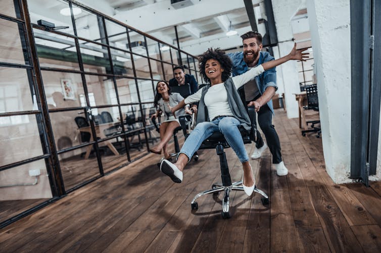 In a modern office, two young men push two young women in rolling office chairs, all are having fun and wearing casual clothing