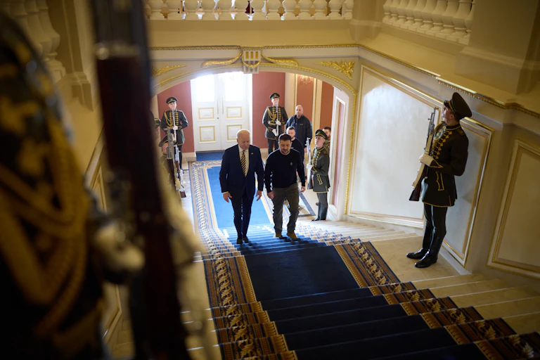 Ukrainian president, Volodymyr Zelensky, and US president, Joe Biden, walk up a carpeted flight of stairs flanked by Ukrainian guards.