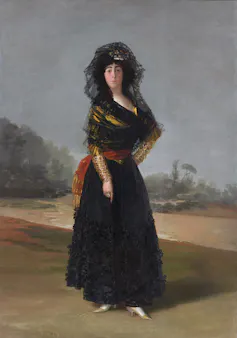 A lone woman stands wrapped in black shawls. She has dark hair and eyebrows and light skin. The backdrop is desolate.