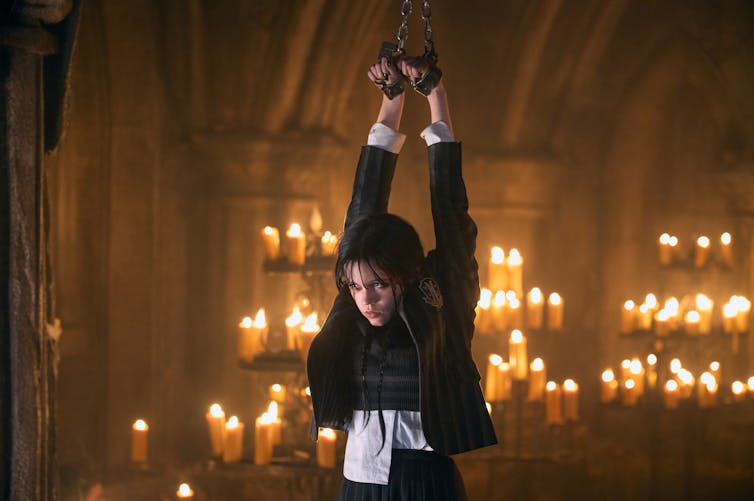 Wednesday Addams is tied up with her arms above her head. Behind her candles burn.