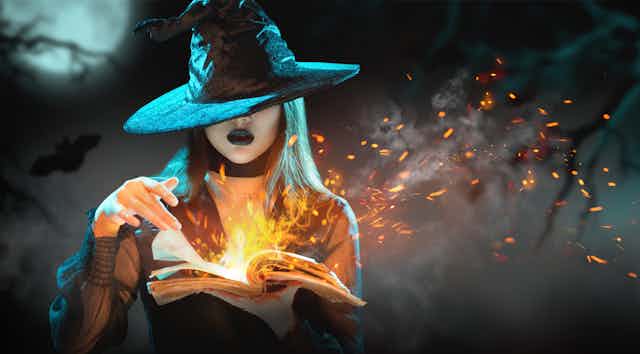 A witch with black hair, a witch hat and black lipstick turns the pages of a flaming book.
