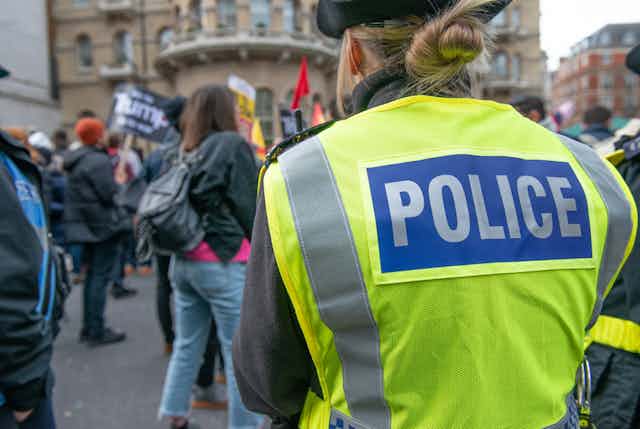 View from behind of a policewoman at a march, with her blonde hair in a bun, wearing a hi-vis vest that reads POLICE