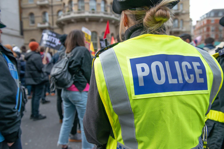 View from behind of a policewoman at a march, with her blonde hair in a bun, wearing a hi-vis vest that reads POLICE