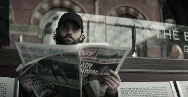 Penn Badgley sits on a bench wearing a cap and reading a newspaper.