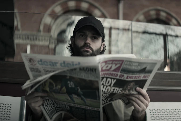 Penn Badgley sits on a bench wearing a cap and reading a newspaper.