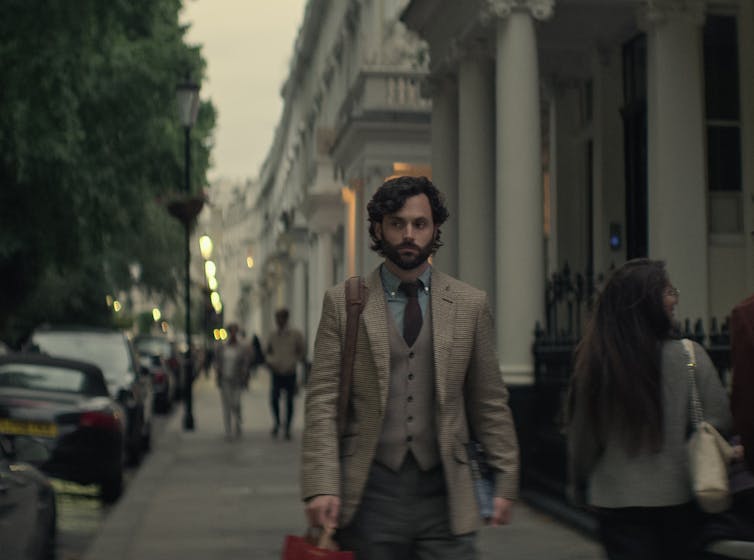 Joe Goldberg wears a tweed suit, walking down a street in Knightsbridge at twilight.