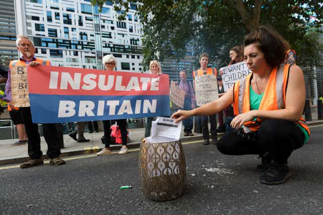People in hi-vis vests stand with an 'Insulate Britain' banner while one burns a letter.