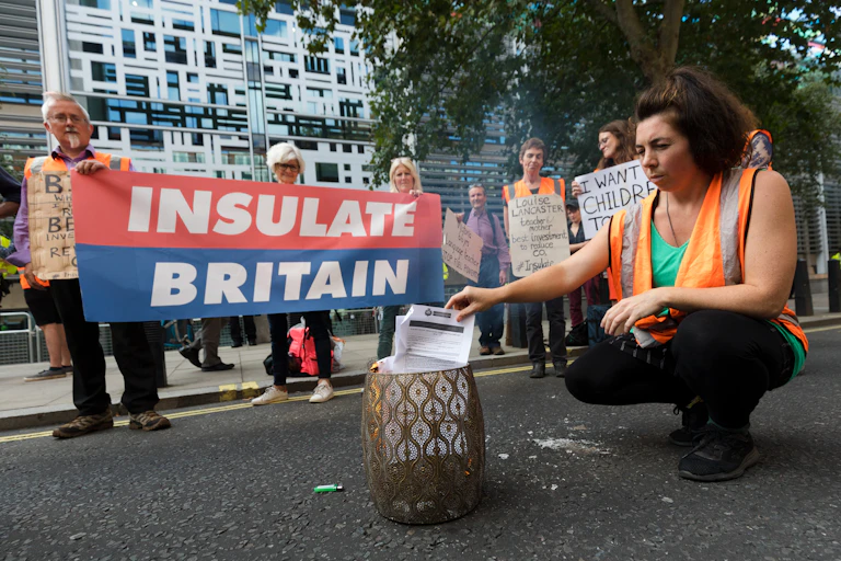 People in hi-vis vests stand with an 'Insulate Britain' banner while one burns a letter.