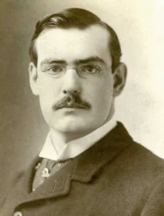 A black and white photo of a young Huntingdon with round glasses a strong moustache and crisp suit.