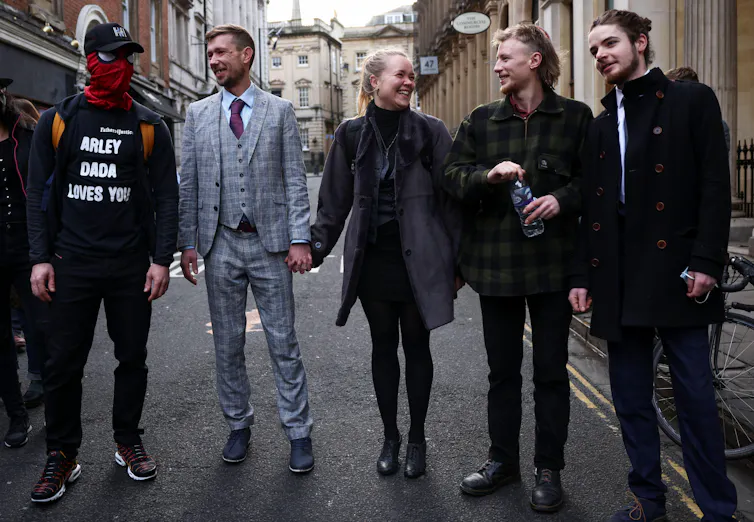Five people in formal attire (and one obscuring their face with a cap and bandana) stand outside a court.