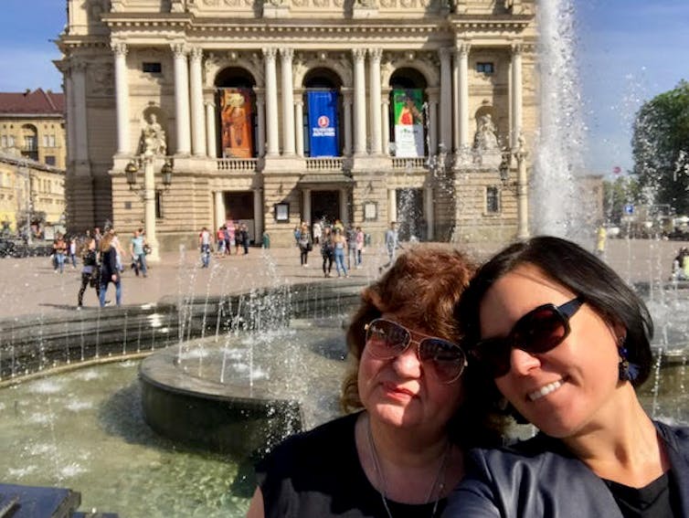 A younger and an older woman, standing in front of a fountain and stately building on a sunny day.