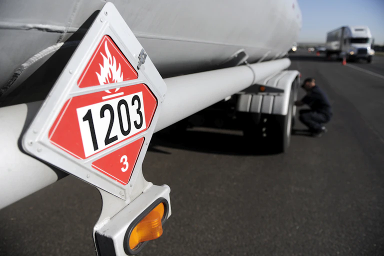 A man squats down beside a truck's tire on a highway as other trucks rush past. The truck he's working on has a placard warming of flammable contents..