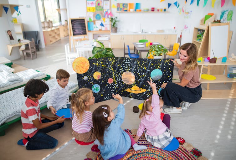 A blond teacher shows young children sitting on the floor a poster about planets.