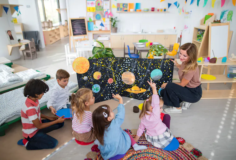 A blond teacher shows young children sitting on the floor a poster about planets.