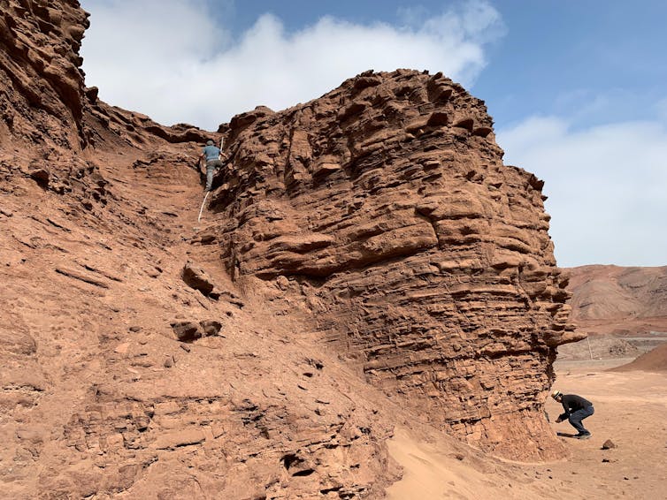 A red, dusty, barren landscape, with a medium size rock formation. One scientist is climing the rock. Another is crouched at its base.