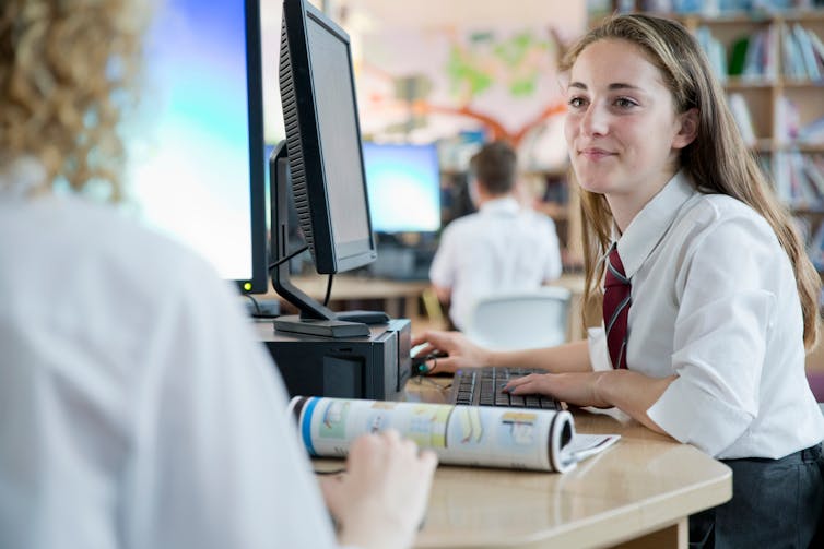 A student works at a computer and smiles at another student.
