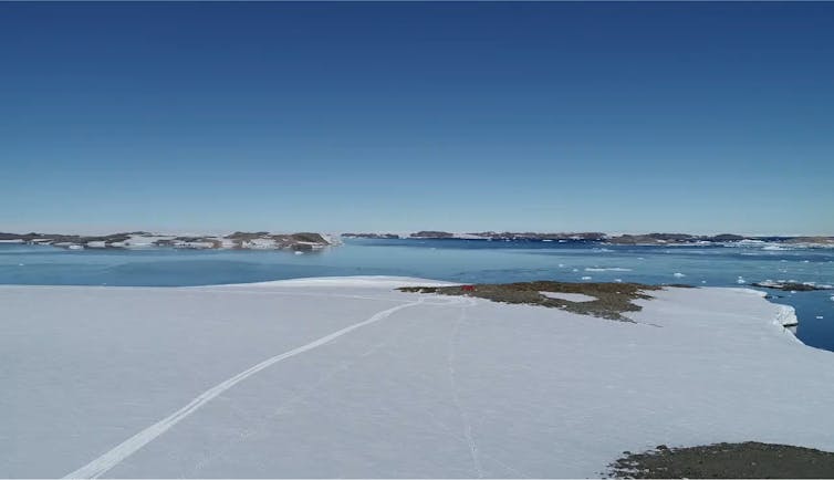 A photo of the vast, barren Antarctic landscape. There is a cloudless blue sky, ice, ocean in the far distance, and a very tiny hut visible in the mid distance.