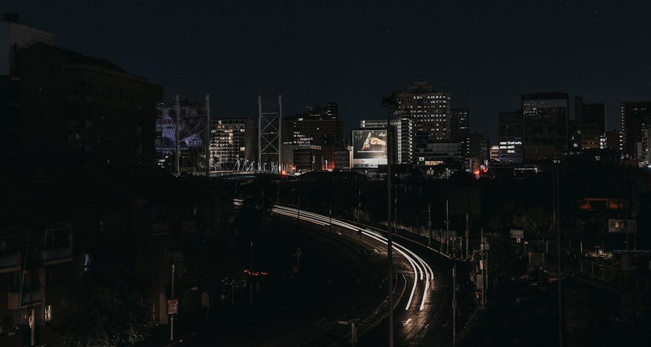 A cityscape at night, with only some buildings and parts of a highway lit up while the other parts are in darkness