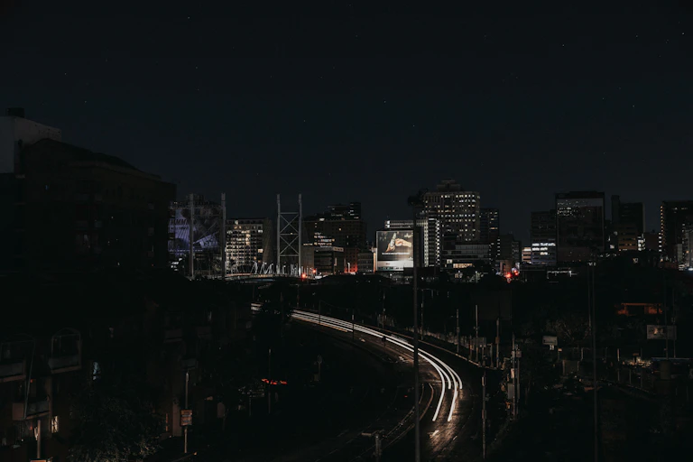 A cityscape at night, with only some buildings and parts of a highway lit up while the other parts are in darkness