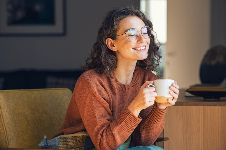A young woman sitting on a couch smiles while holding a mug.