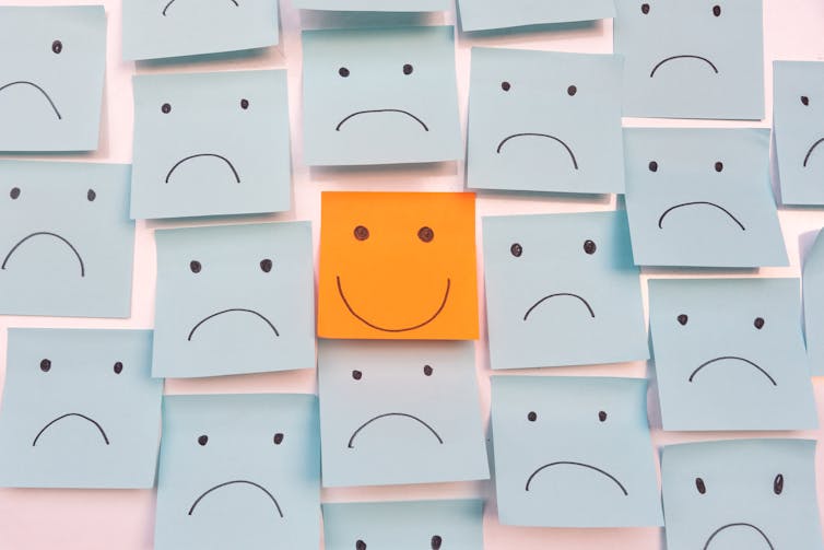 A wall of blue sticky notes with frowns on it. In the centre is an orange sticky note with a smiley face on it.