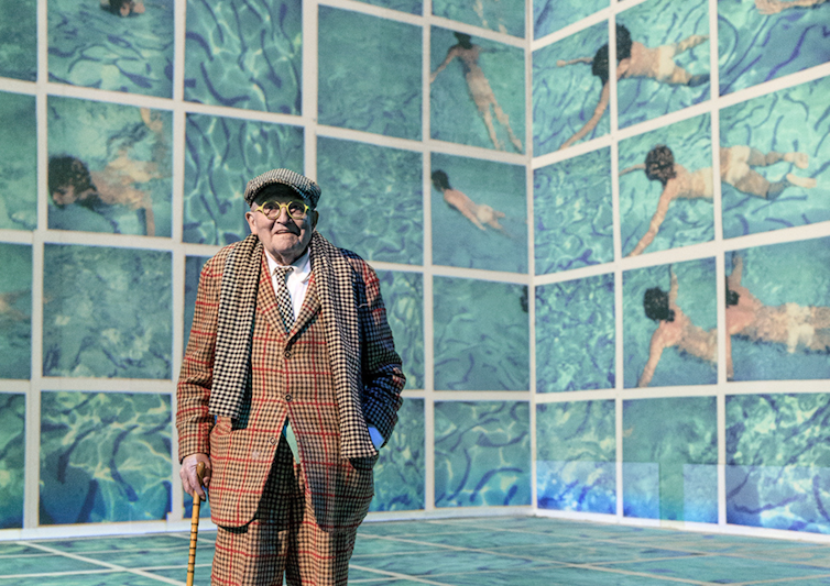 An old man in a tweed suit stands in front a screen with a painting of swimmers on it.